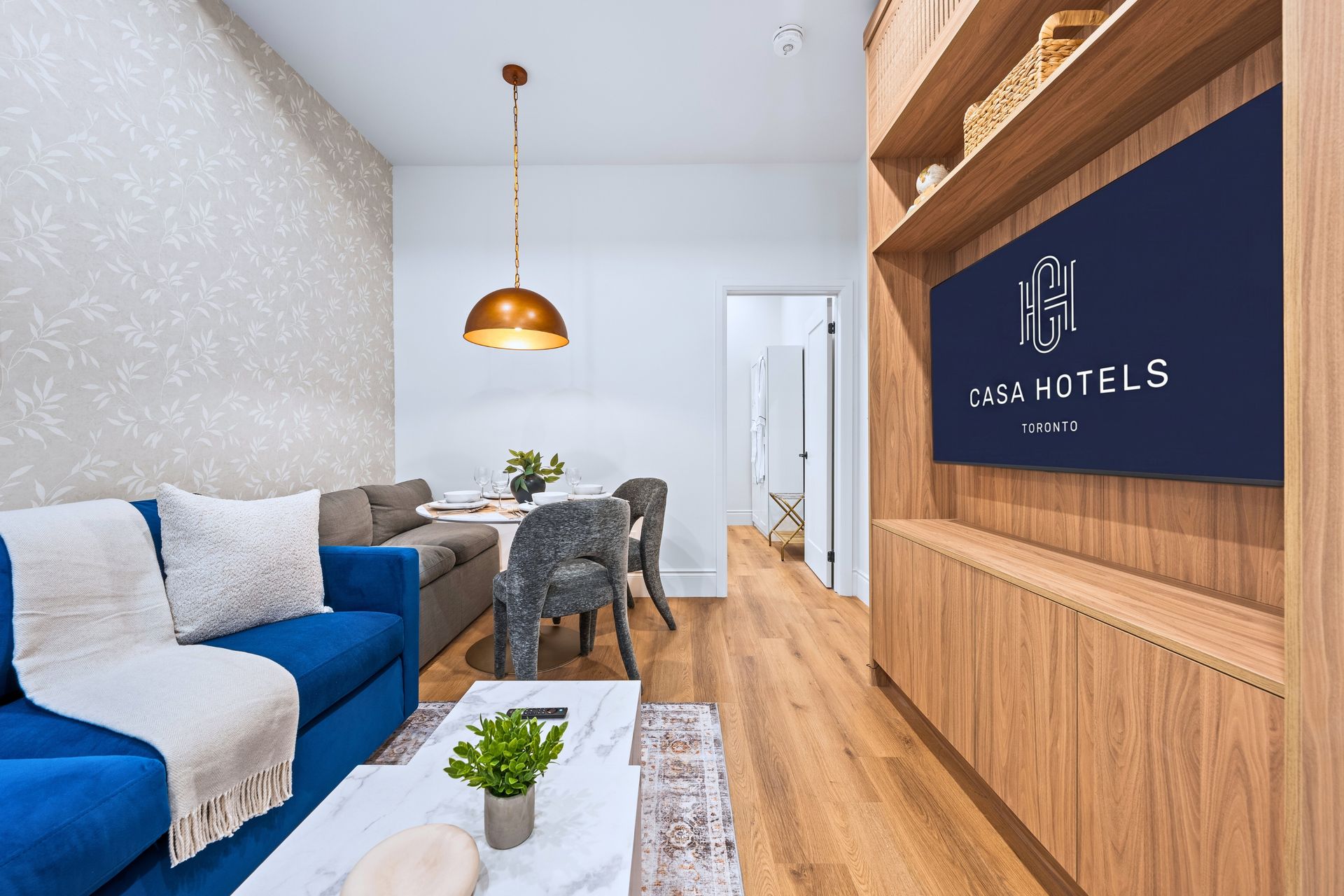Modern living room with blue sofa, gray chairs, wood floor, pendant light, and wall-mounted TV