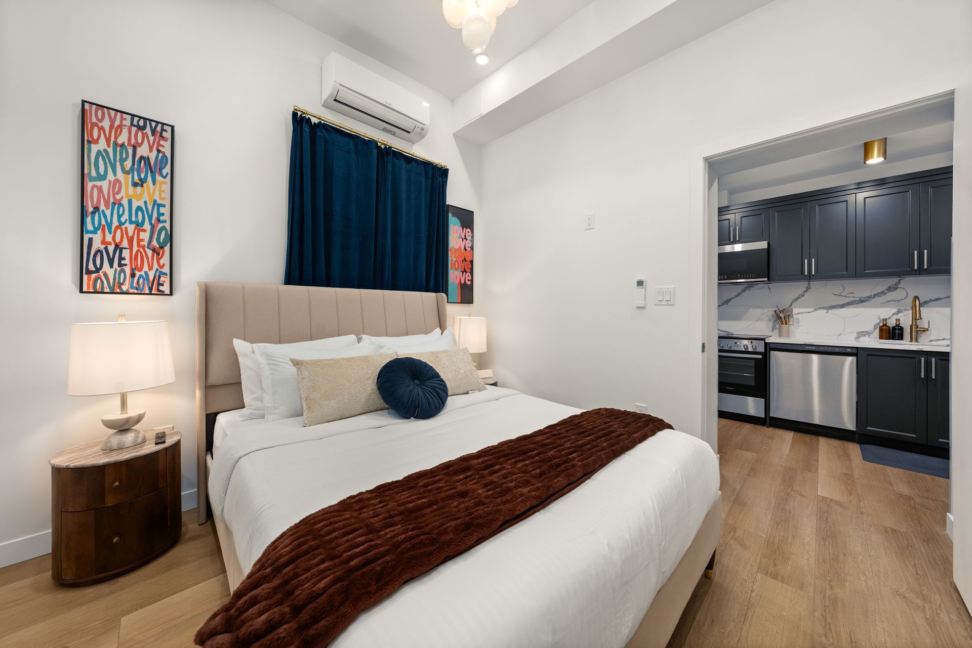 Bedroom with a bed, artwork, and open doorway to a kitchen with dark cabinets and appliances.