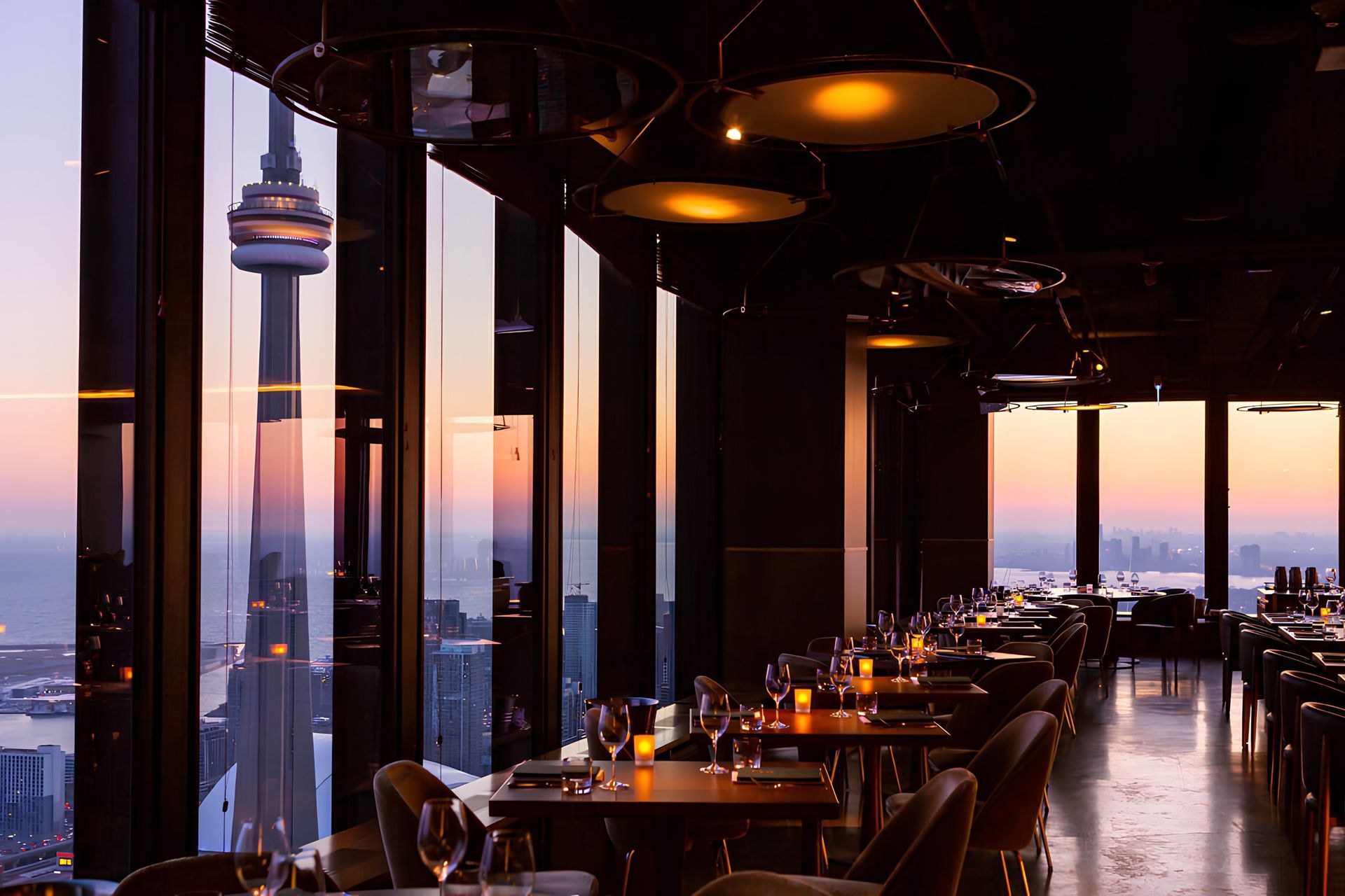 Panoramic restaurant with floor to ceiling windows overlooking the Toronto skyline at sunset, warmly lit tables inside
