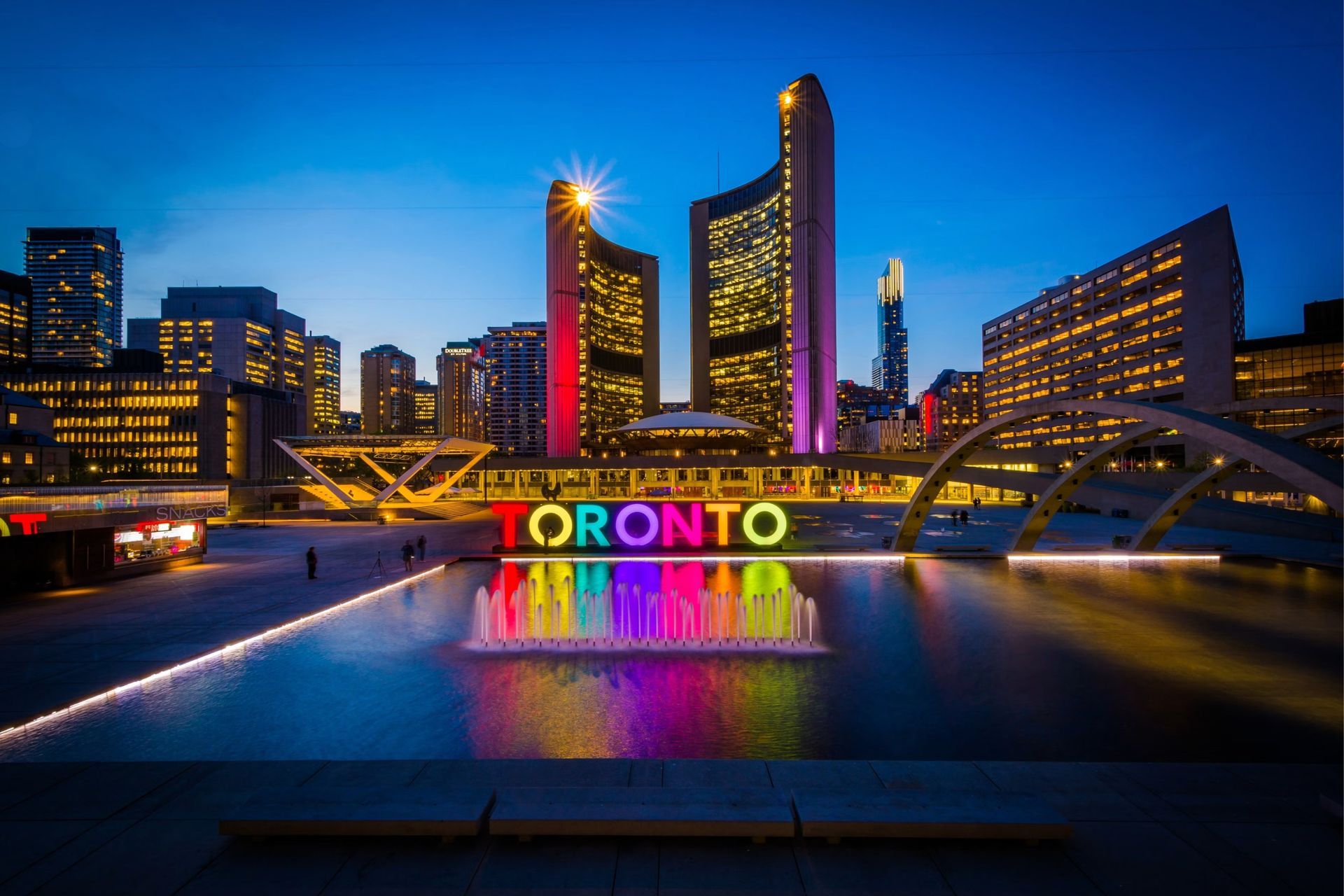 Toronto skyline at dusk with illuminated buildings and colorful reflected lights over the water.