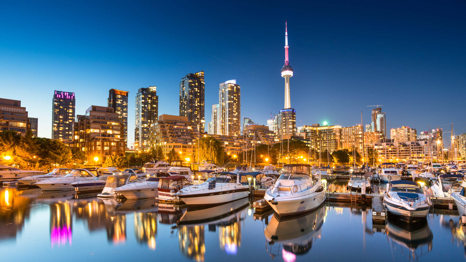 Toronto skyline and marina at dusk, with boats reflecting colorful city lights and the CN Tower in the distance
