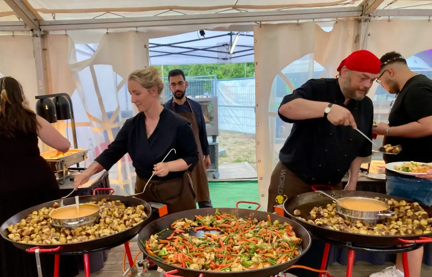 Eine Gruppe von Menschen kocht in einem Zelt Essen in großen Pfannen.