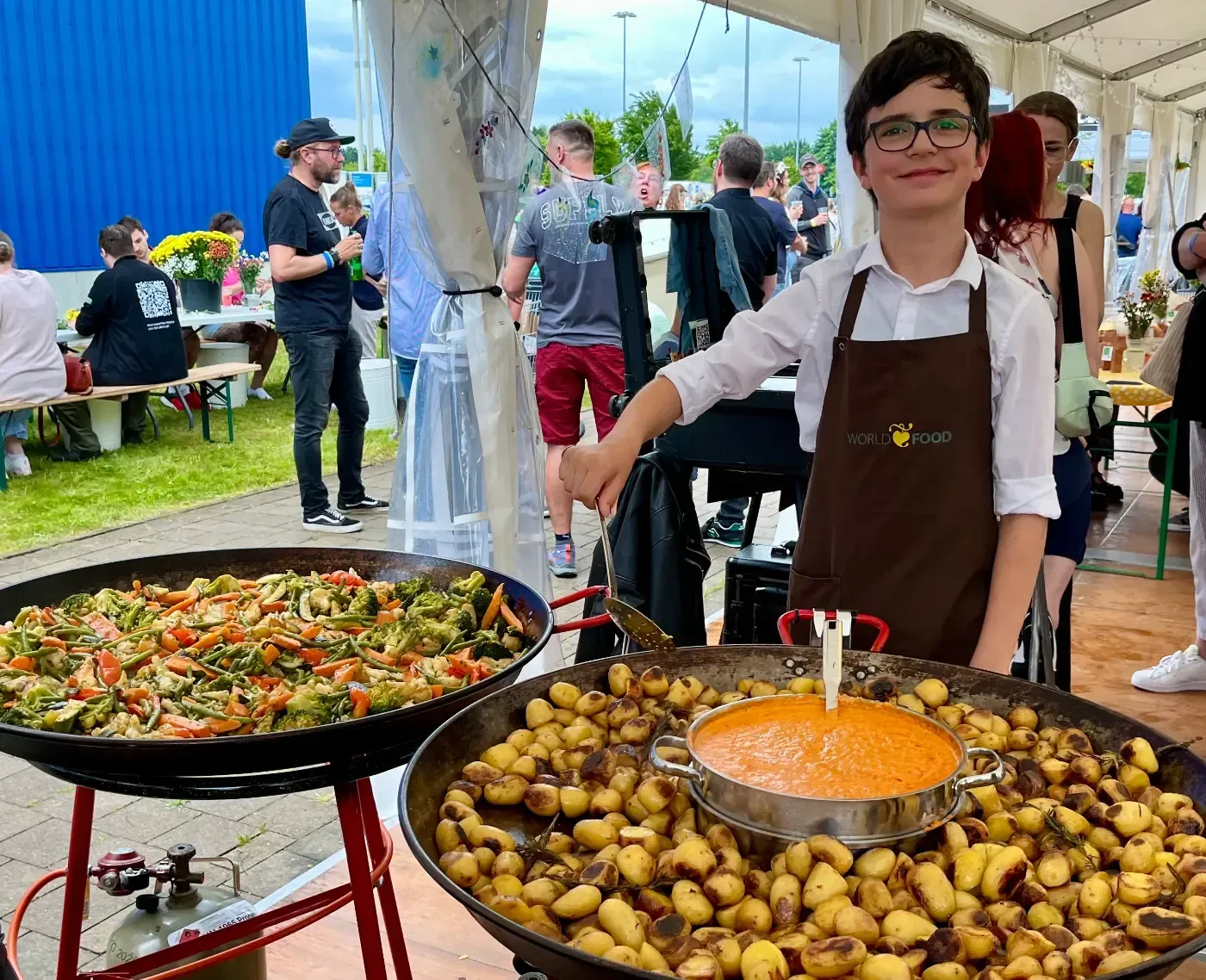 Ein Junge in einer Schürze steht vor zwei Pfannen mit Essen.