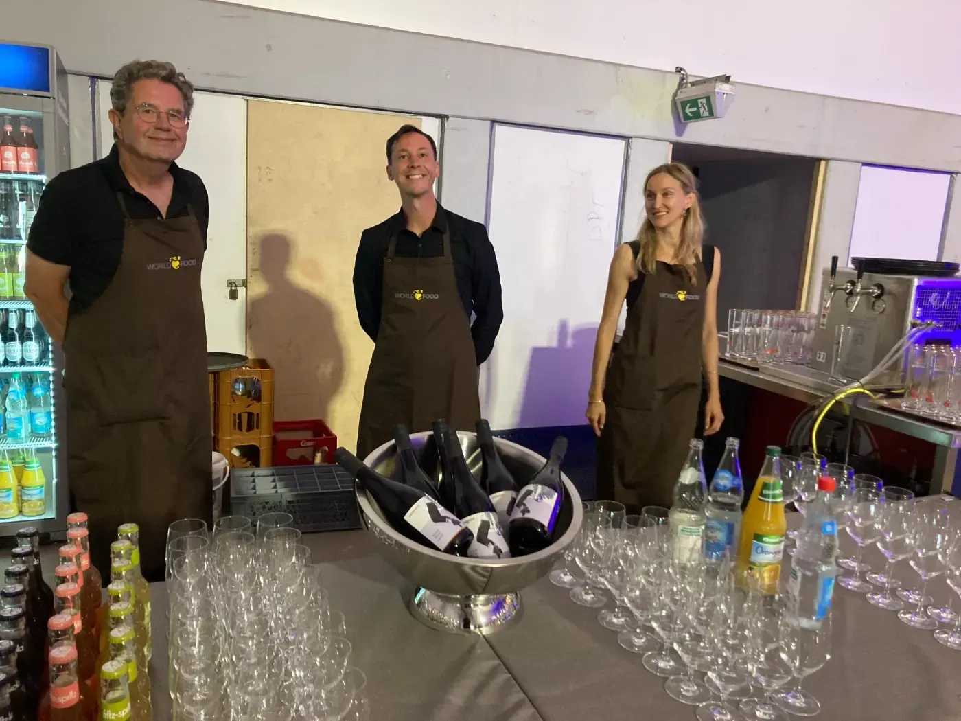 Eine Gruppe von Menschen steht mit Weinflaschen und Gläsern um einen Tisch herum.