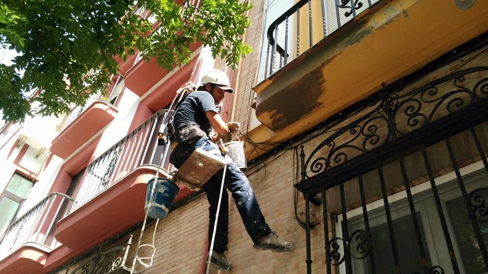 Obrero con cuerdas, reparando la fachada de un edificio. Lleva arnés y casco, frente a un edificio de estilo español.