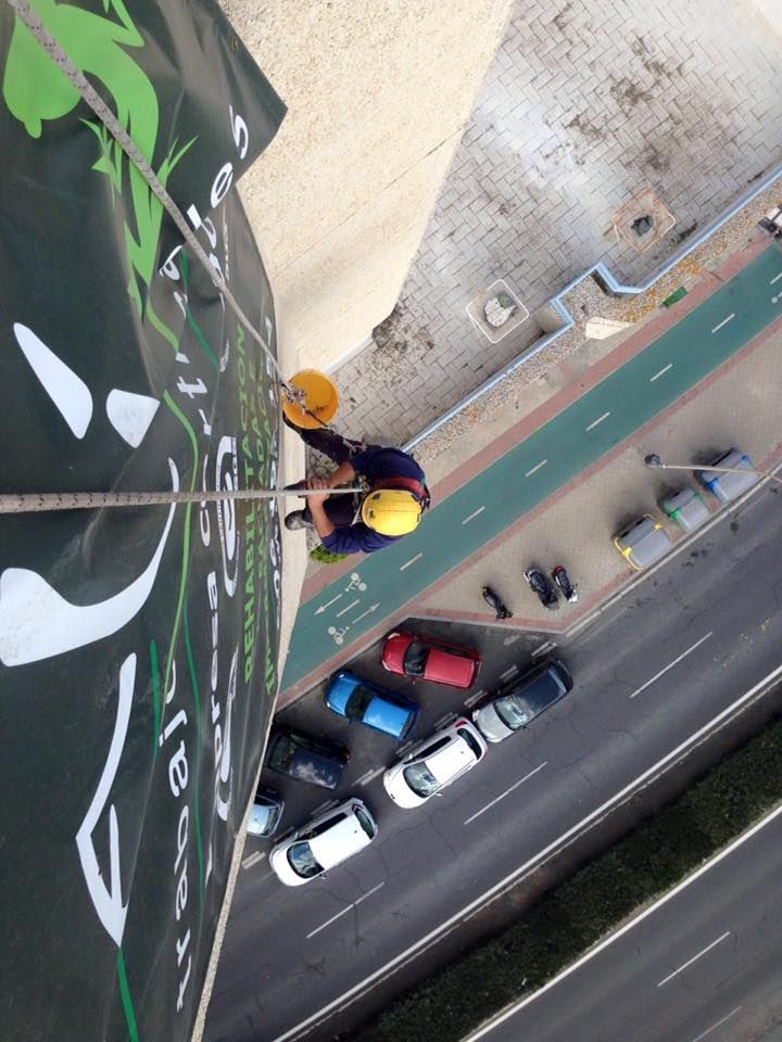 Una persona desciende en rápel por un edificio alto; hay una señalización verde, coches debajo y un casco amarillo.