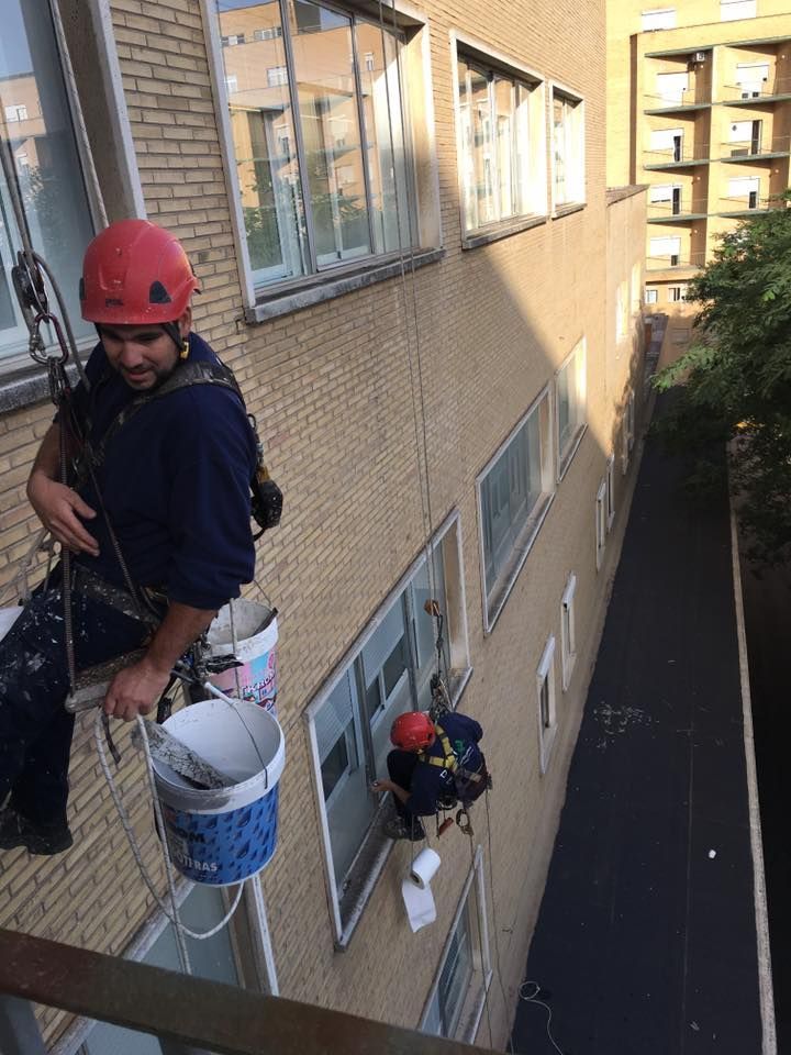 Dos pintores con cuerdas, trabajando en el exterior de un edificio. Uno sostiene un cubo, el otro usa una herramienta.