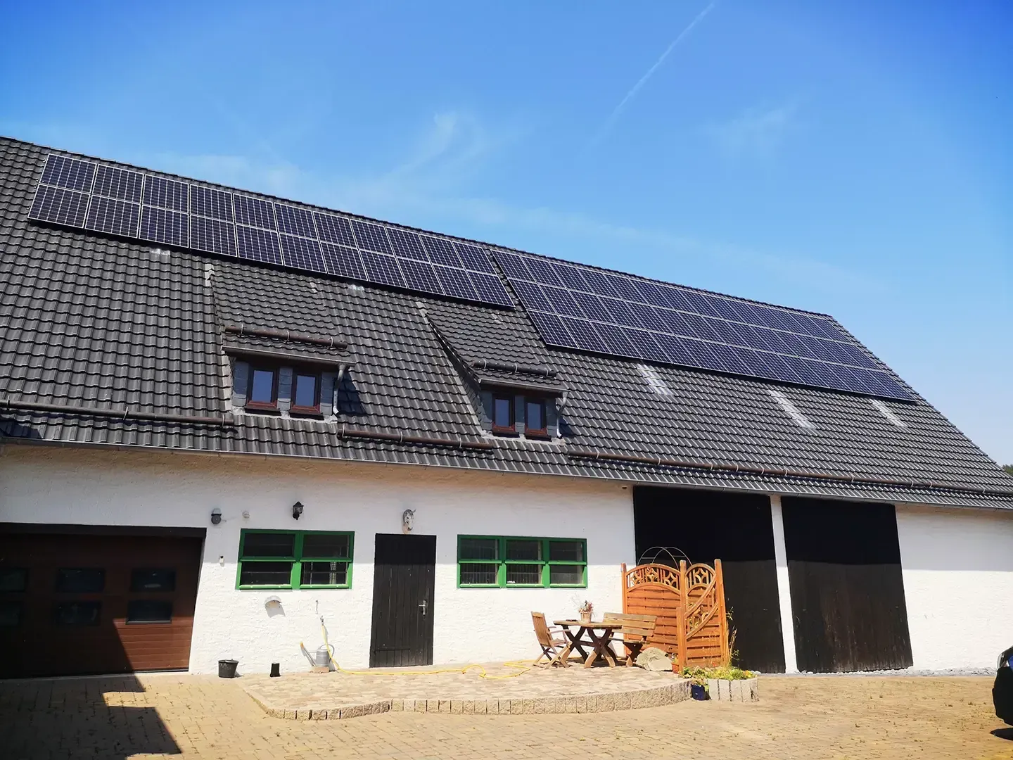 Eine Scheune mit Solarpaneelen auf dem Dach, weißen Wänden und grünen Fensterrahmen unter blauem Himmel.
