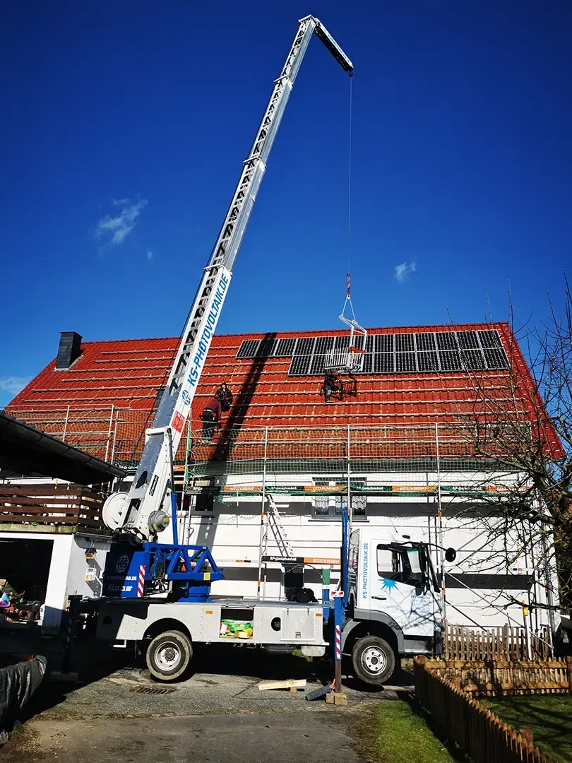 Ein LKW-Kran hebt Baumaterialien auf ein Hausdach. Im Hintergrund rote Ziegel und blauer Himmel.
