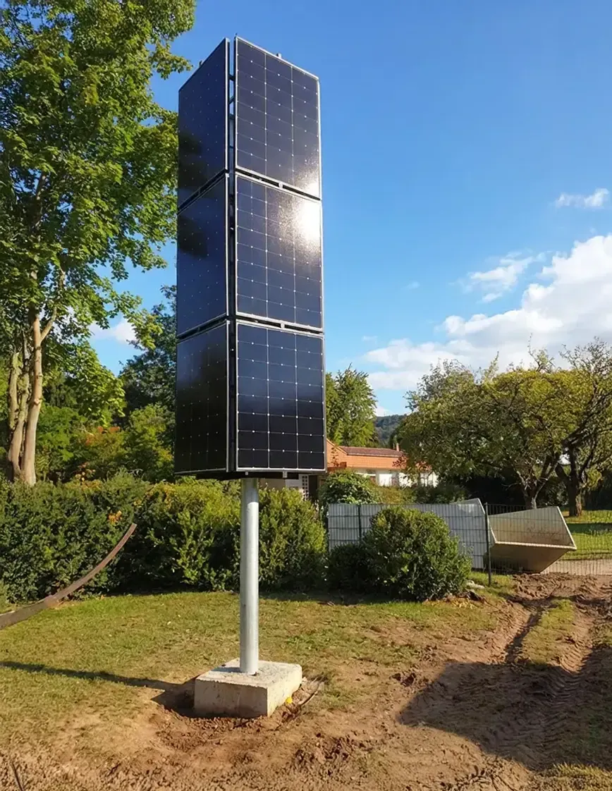 Eine hohe, vertikale Solarpanelanlage auf einem Mast in einem grasbewachsenen Garten unter blauem Himmel.