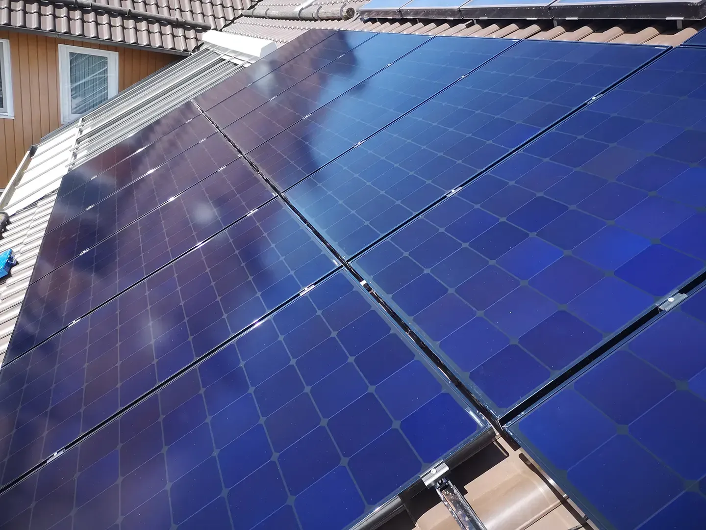 Auf einem Dach installierte Solarpaneele. Blaue Paneele mit reflektierender Oberfläche vor einem braunen Dach und weißer Fassadenverkleidung.