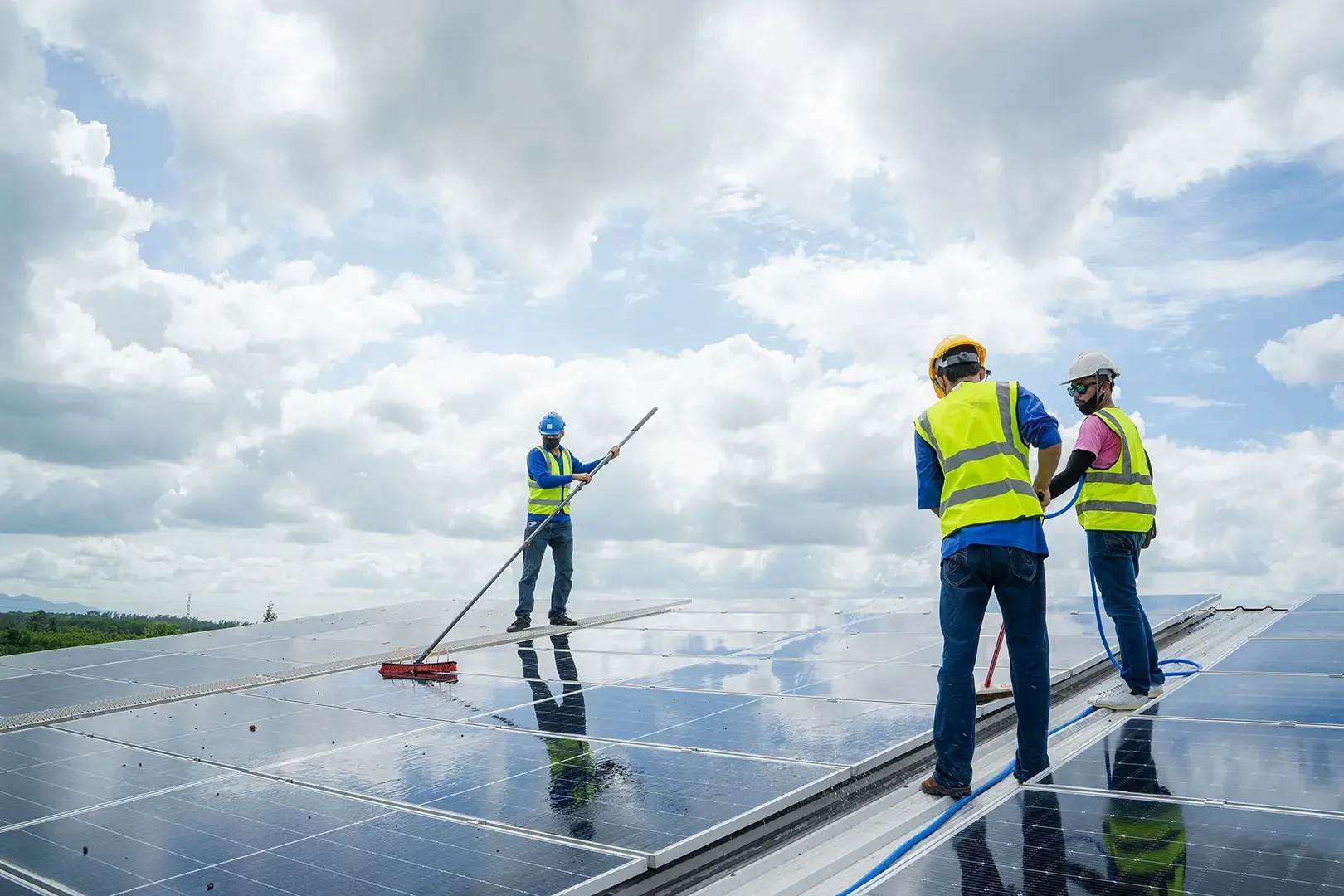 Arbeiter in Sicherheitswesten reinigen Solarpaneele auf einem Dach unter bewölktem Himmel.