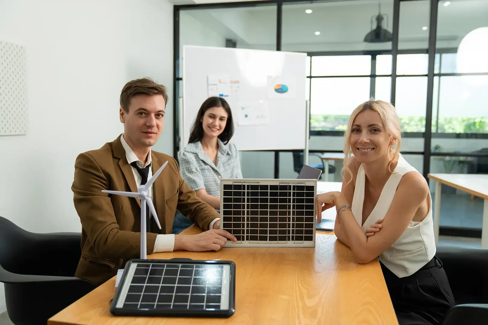 Drei Personen in einem Besprechungsraum mit Modellen von Solarmodulen und Windkraftanlagen. Sie lächeln in die Kamera.