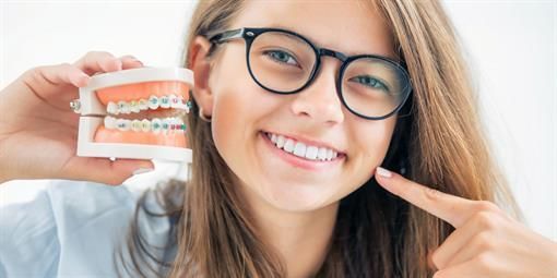 Ein junges Mädchen mit Brille hält ein Zahnmodell und zeigt auf ihr Lächeln.