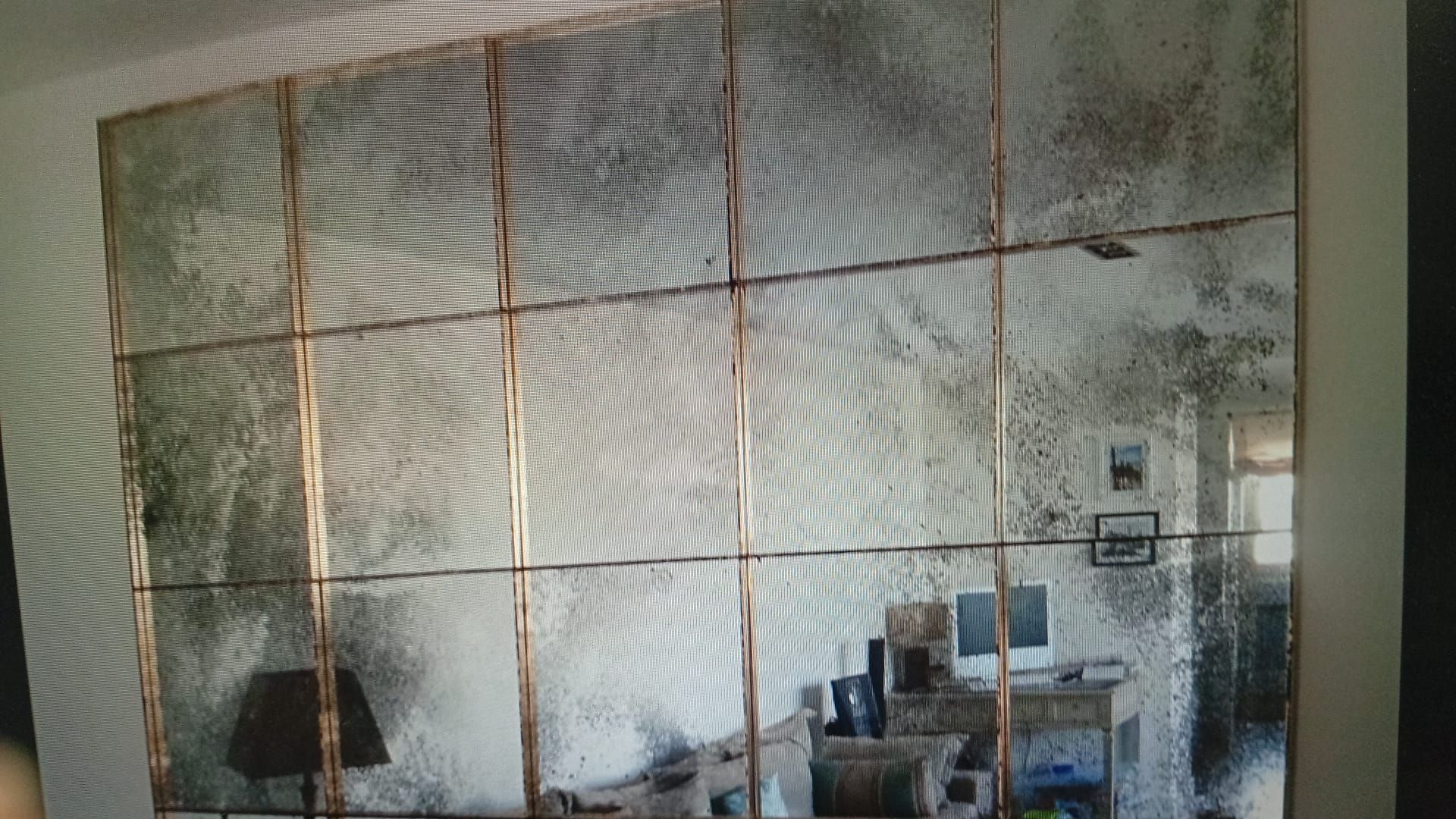 Gran espejo de pared de estilo antiguo con diseño de cuadrícula, que refleja una habitación con muebles y ventana.