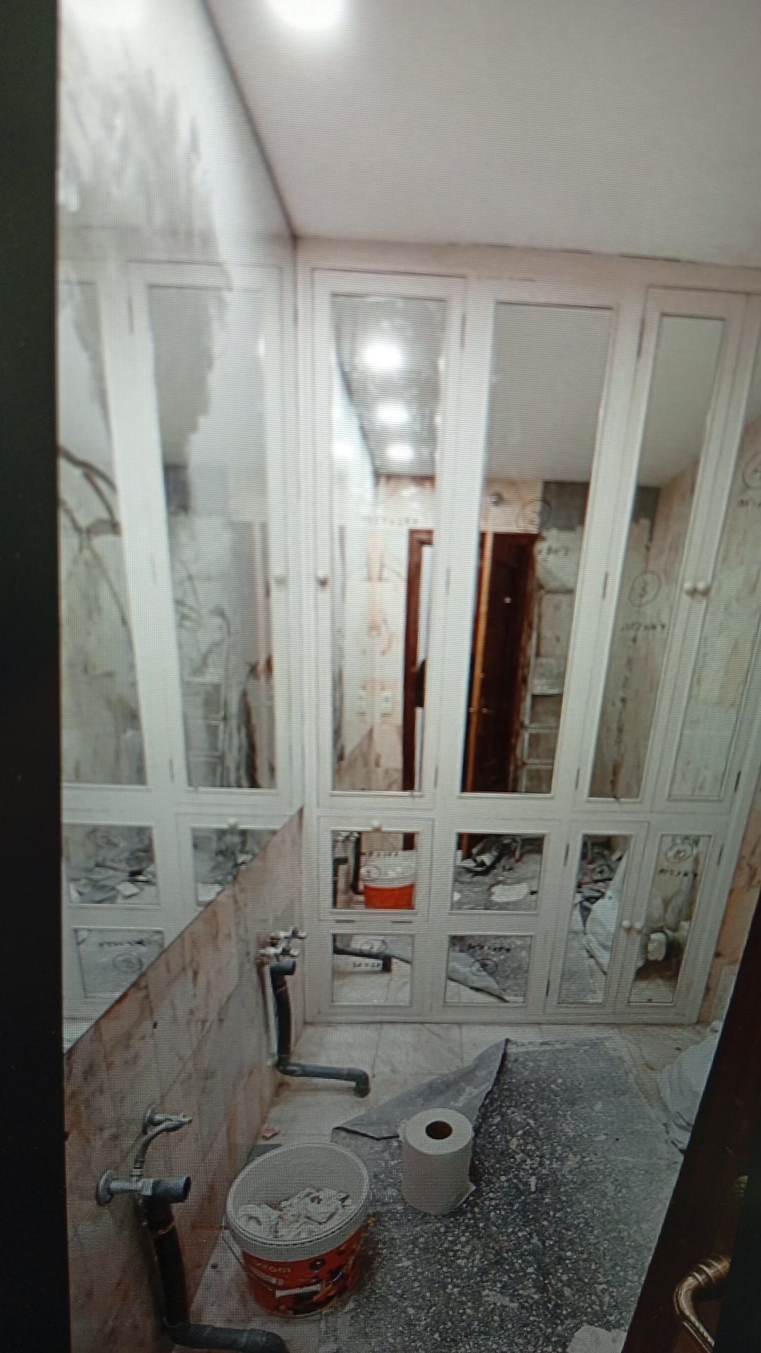 Baño en construcción con ventanas con marco blanco y un balde en el suelo.