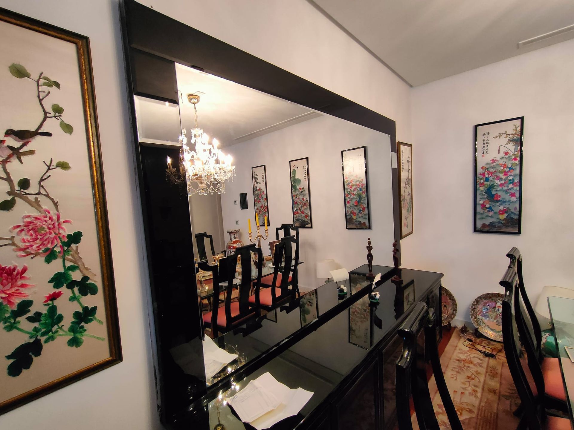 Un gran espejo negro refleja un comedor con una lámpara de araña, obras de arte y un aparador.