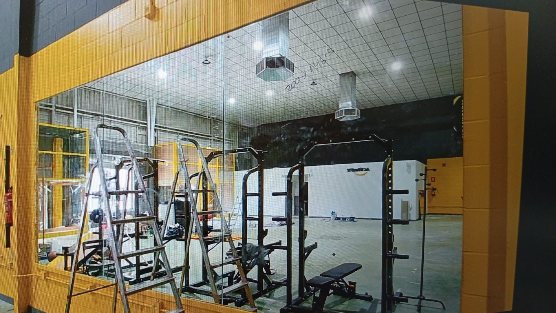 Espejo que refleja un gimnasio con marco amarillo y equipamiento: estante de pesas, banco y una escalera, ubicados en un gran espacio abierto.