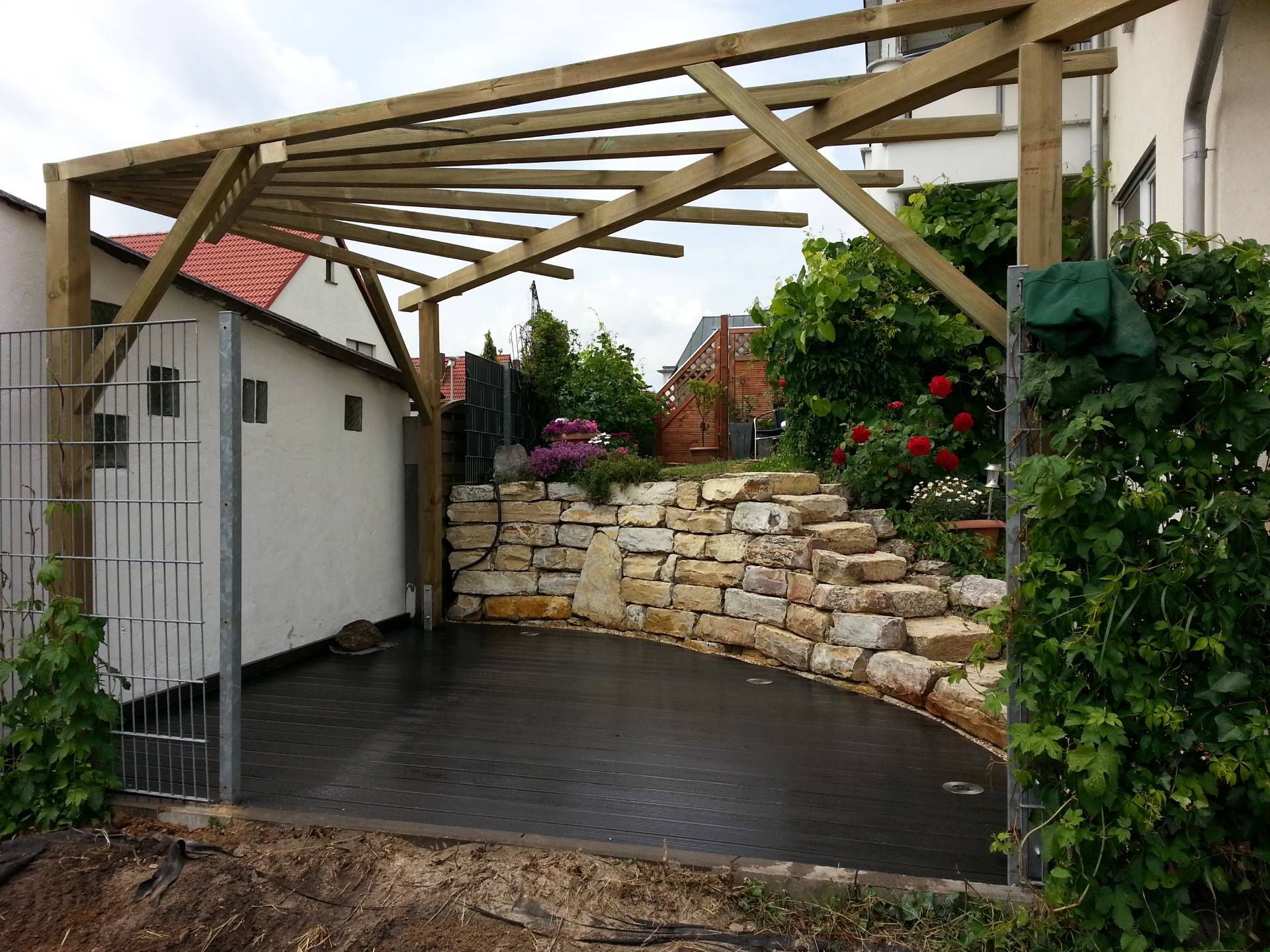 Holzpergola mit offenem Dach über dunklem Bodenbelag, davor eine niedrige Mauer aus Natursteinen mit Pflanzen und Blumen.