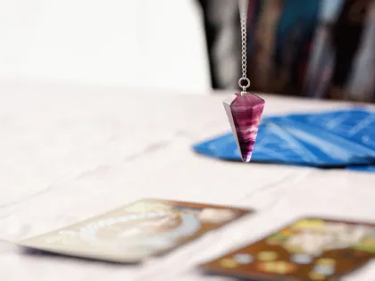 El péndulo cuelga sobre cartas del tarot sobre una superficie blanca; tela azul y figuras borrosas en el fondo.