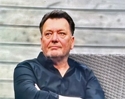 Ein Mann mit Bart sitzt mit verschränkten Armen auf einem Stuhl.