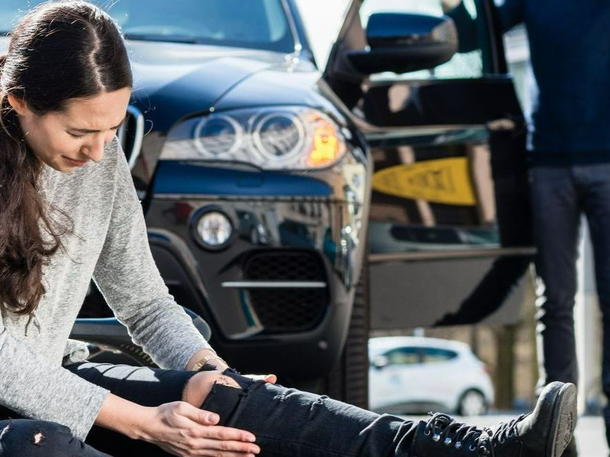 Une jeune femme par terre qui tient son genou et une voiture en arrière-plan.