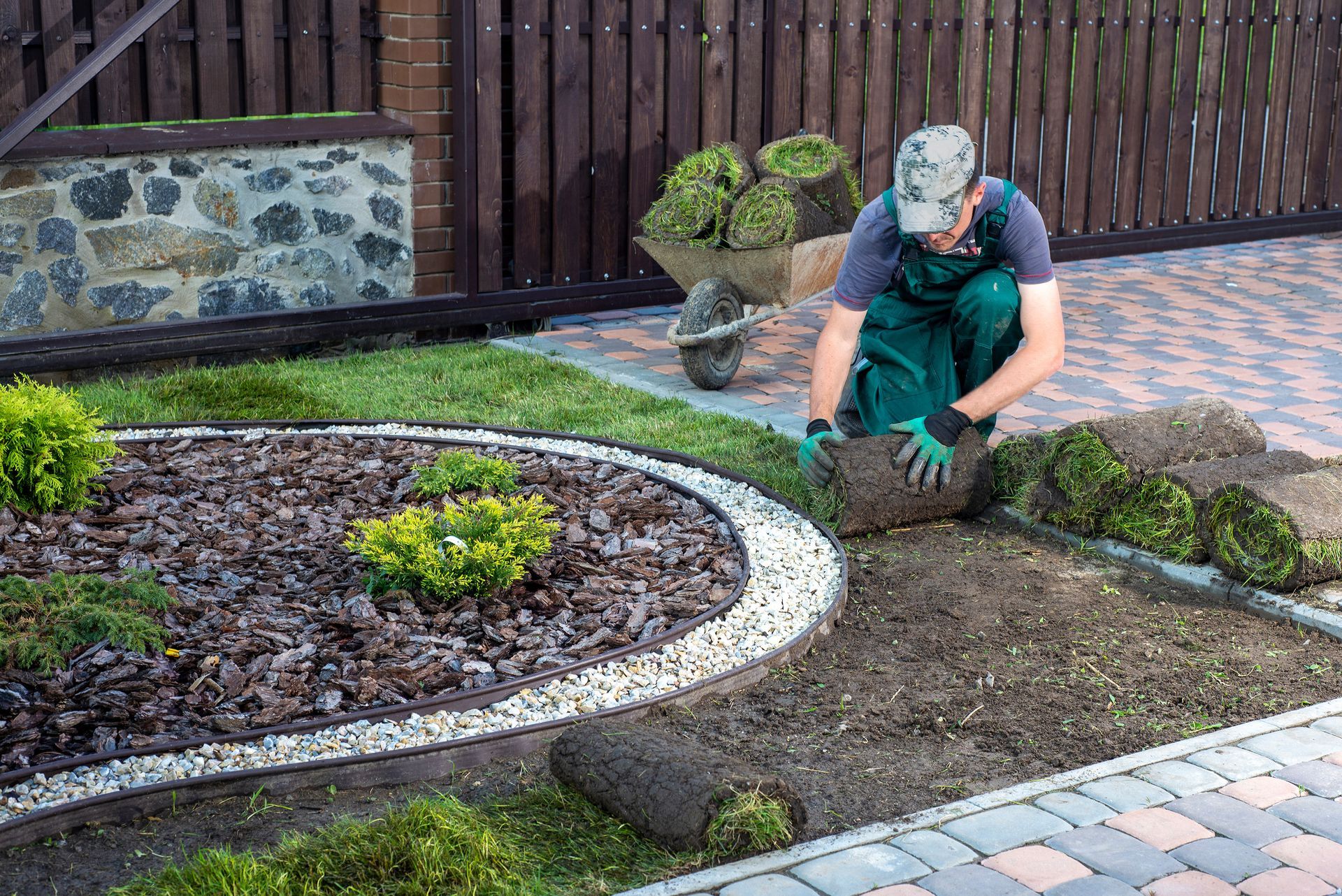 Un ouvrier coiffé d'une casquette et vêtu d'une salopette verte installe du gazon en rouleaux dans un jardin, le long d'un parterre de paillis bordé de pierres.
