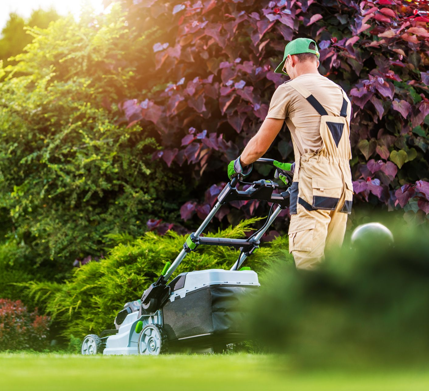 Une personne en uniforme beige et casquette verte pousse une tondeuse à gazon sur une pelouse herbeuse, devant des buissons de jardin luxuriants.