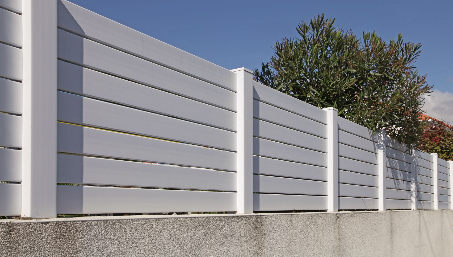 Une clôture blanche horizontale à lattes régulièrement espacées, fixée sur un mur de béton gris, se détachant sur un ciel bleu clair.