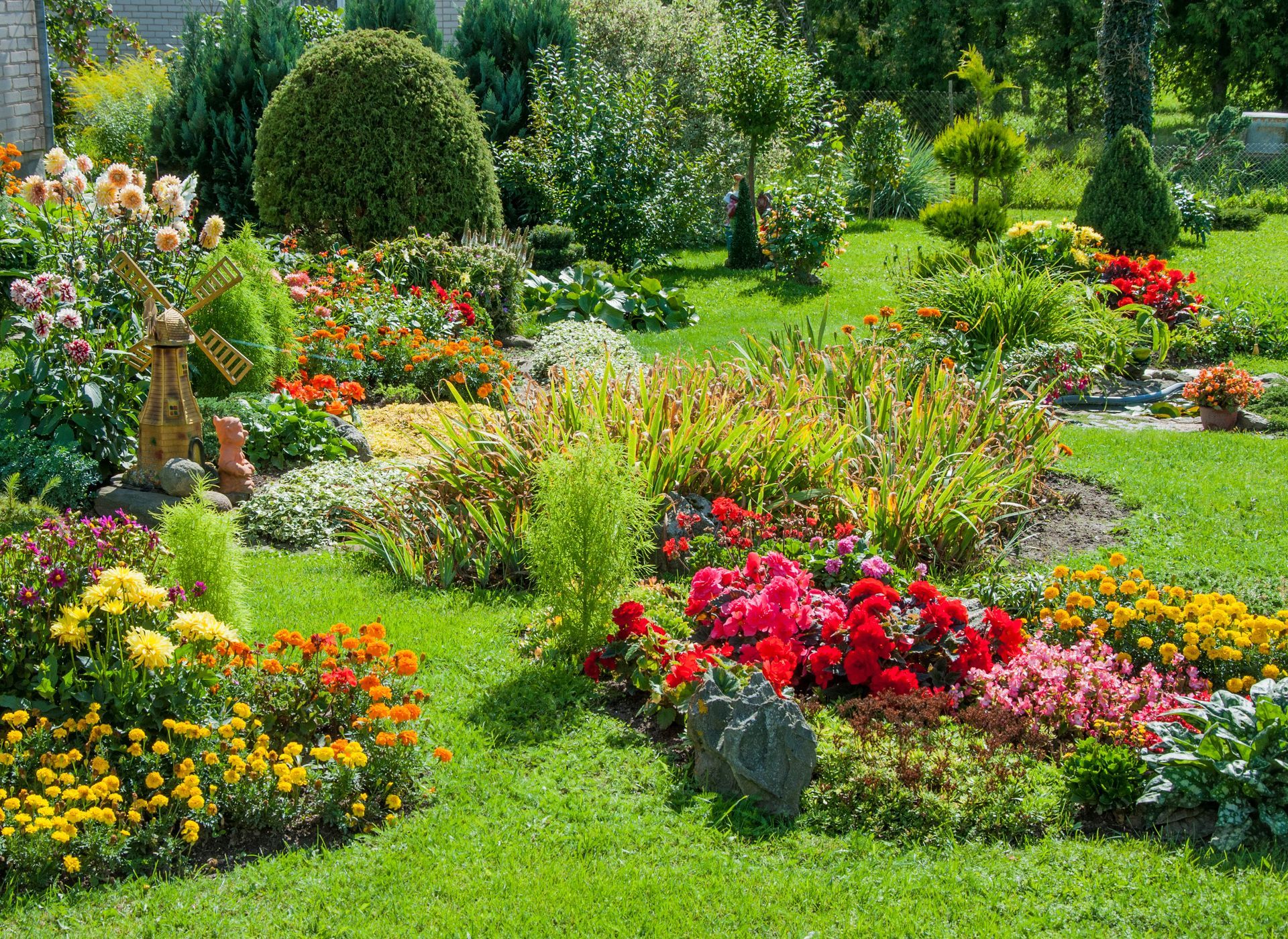 Un jardin luxuriant agrémenté d'un moulin à vent décoratif, de parterres de fleurs aux couleurs éclatantes (rouge, jaune et orange) et d'une végétation soignée.