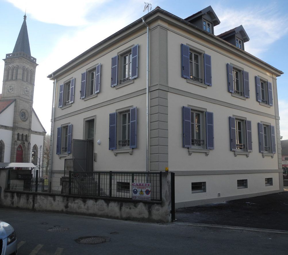 Bâtiment beige de deux étages avec des volets bleus, à côté d'une église avec un clocher, dans une rue.