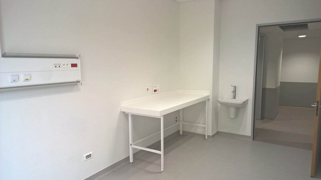 Une salle d'examen médical aux murs blancs, avec une table, un lavabo et une porte ouverte donnant sur une autre pièce.
