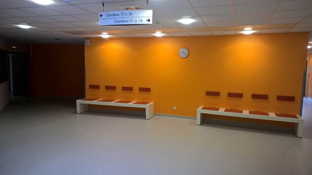 Mur orange avec bancs et horloge dans une salle d'attente. Panneau de signalisation au-dessus.