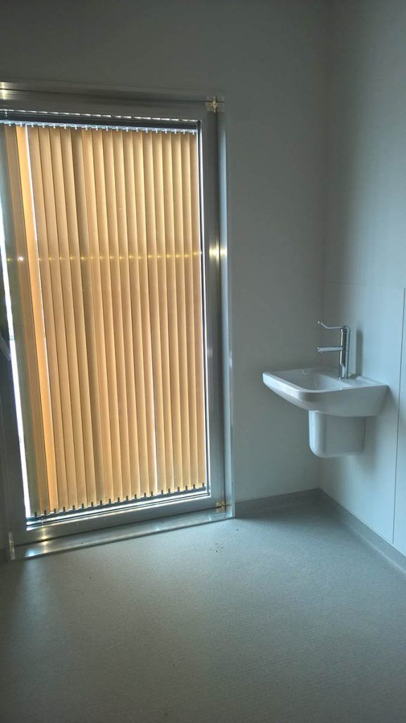 Une petite pièce avec une fenêtre recouverte de stores verticaux, un lavabo mural et un sol gris.