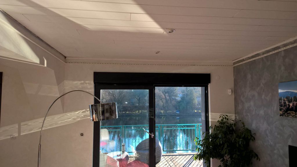 Intérieur d'une chambre avec vue sur un lac à travers une porte coulissante en verre. Plafond et murs baignés de lumière naturelle, plante dans un coin.