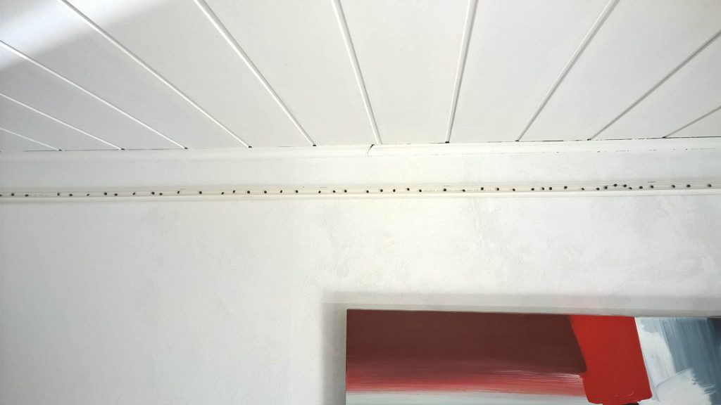 Plafond blanc lambrissé, au-dessus d'un mur blanc percé d'une porte donnant sur une autre pièce.