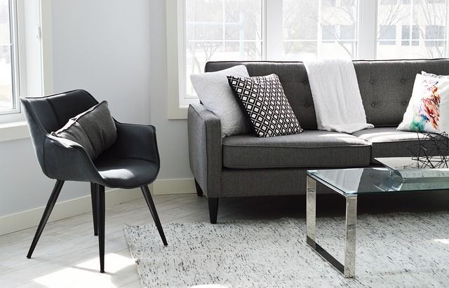 Salon avec canapé gris, fauteuil, tapis et table basse en verre près d'une fenêtre.