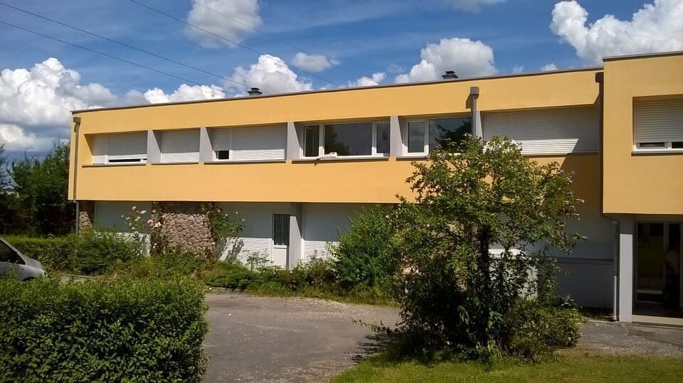 Bâtiment jaune de deux étages avec de nombreuses fenêtres, entouré de buissons verts et d'un ciel bleu nuageux.