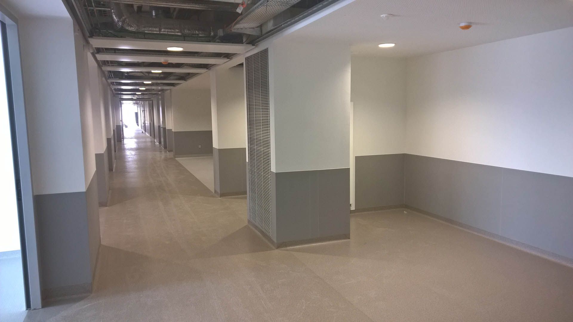 Long couloir aux murs et colonnes gris et blancs ; une personne se profile au loin.