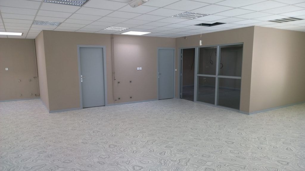 Espace de bureau vide avec murs beiges, portes grises et sol moucheté. Éclairage fluorescent au plafond.