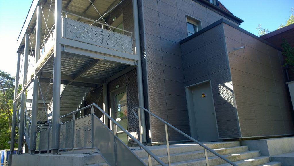 Vue extérieure d'un bâtiment avec un escalier métallique, des marches en béton et un revêtement gris foncé.
