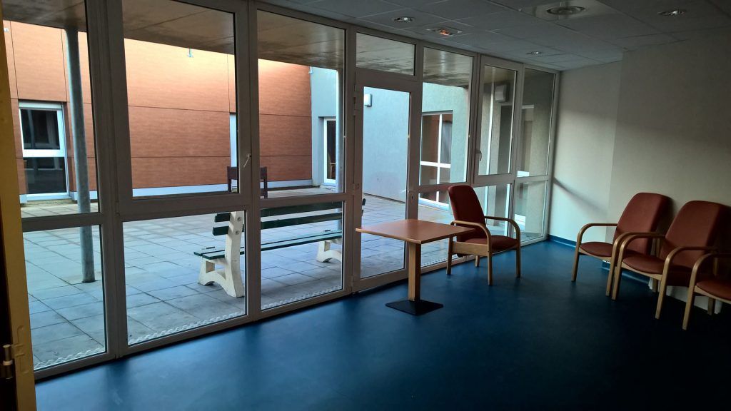 Salle d'attente avec sol bleu, paroi vitrée donnant sur la cour intérieure, chaises et table.