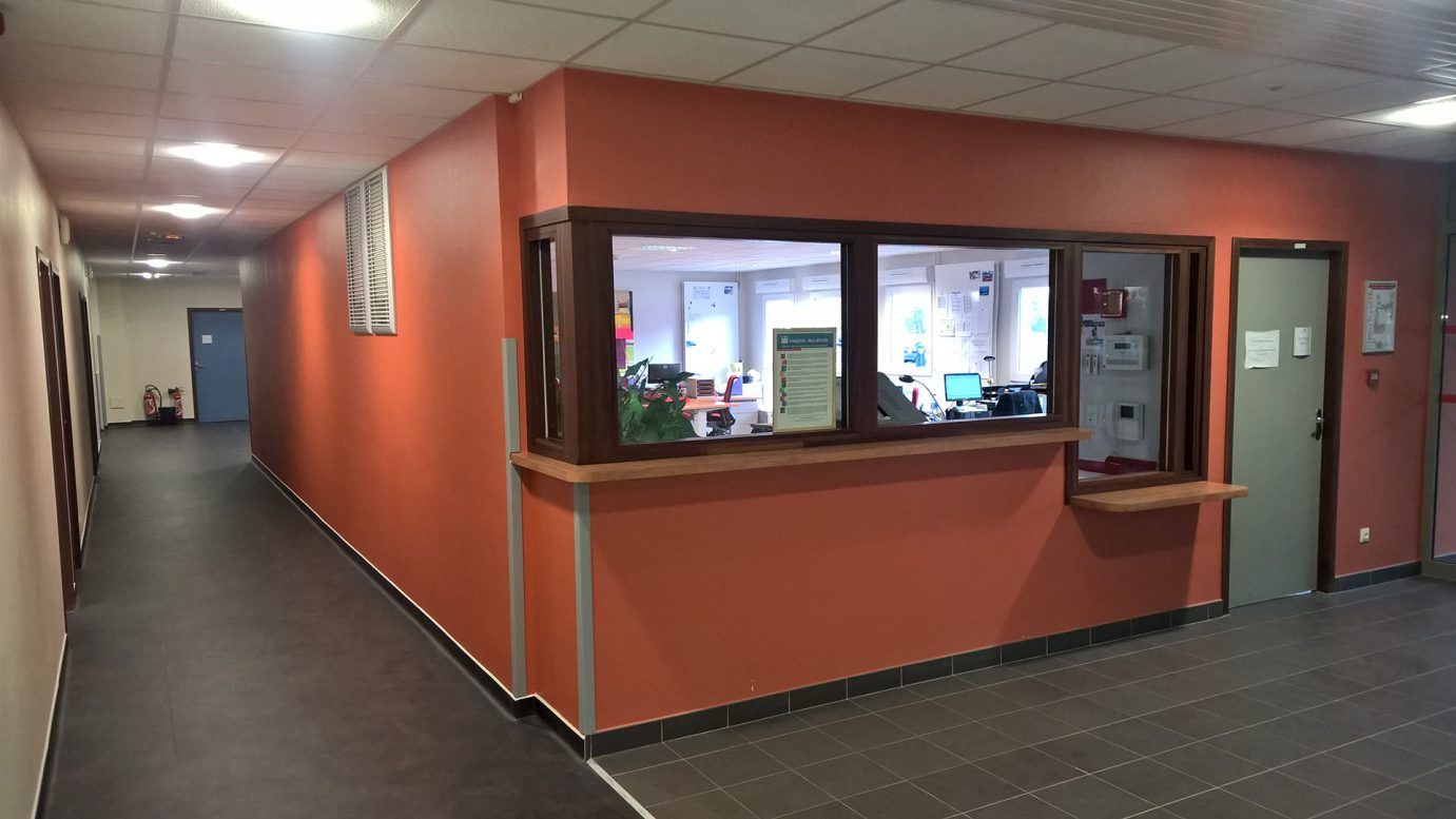 Un couloir menant à un bureau avec un guichet de service intégré dans un mur couleur terre cuite, et un sol carrelé.