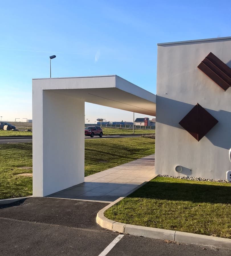 Entrée d'un immeuble moderne blanc avec un abri voiture et des décorations murales en forme de losanges bruns.