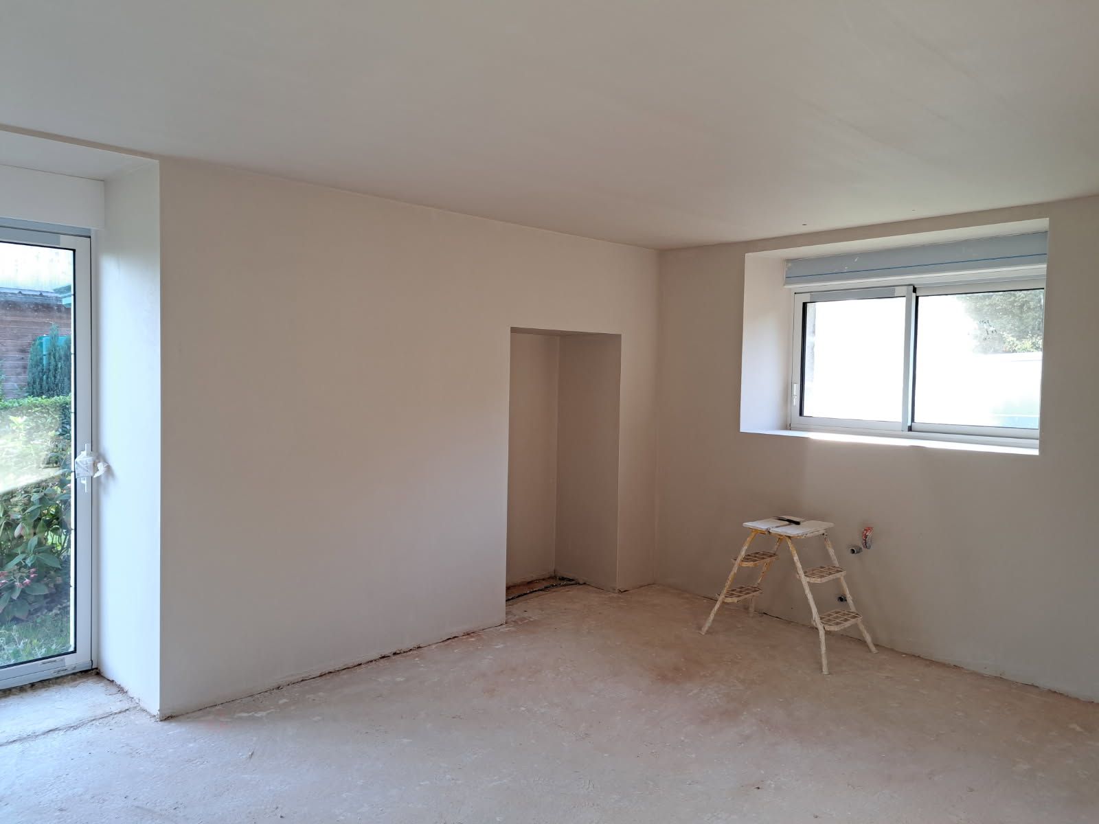 Pièce vide aux murs clairs, une porte, une fenêtre avec des stores et un petit escabeau.