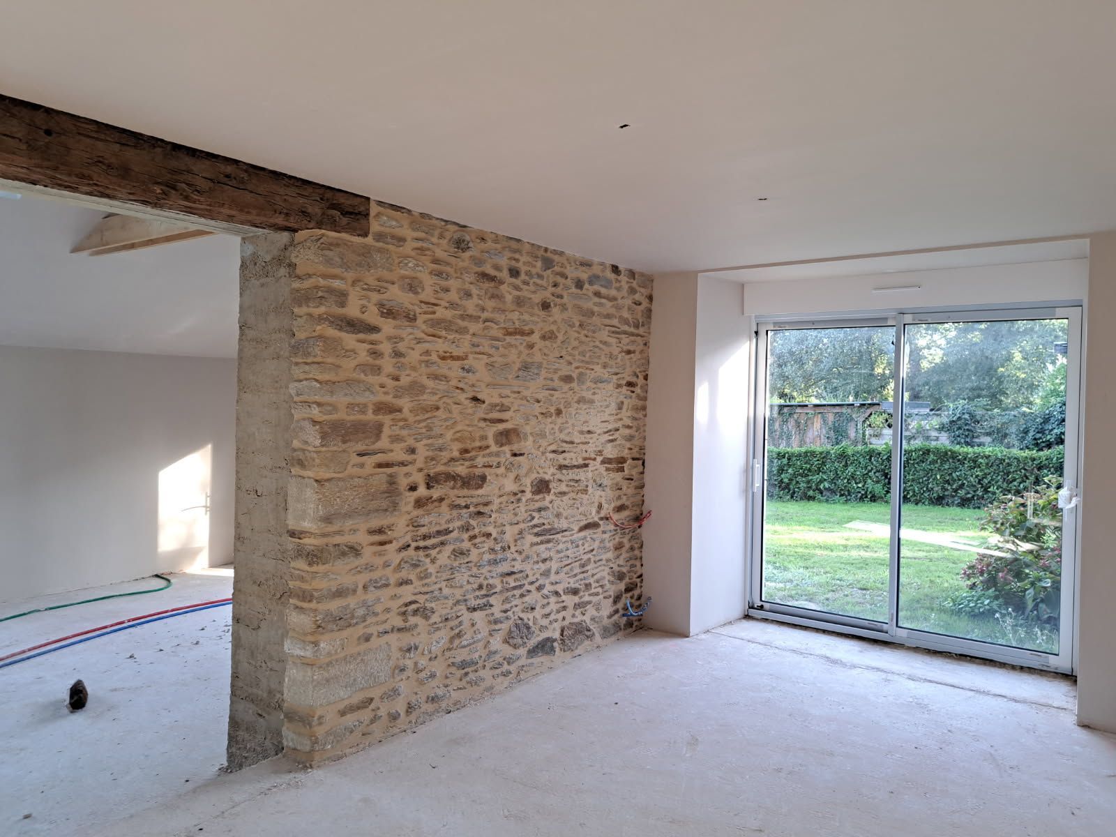 Vue intérieure d'une pièce avec un mur en pierre apparente, une porte coulissante en verre et des murs et un sol en plâtre brut.