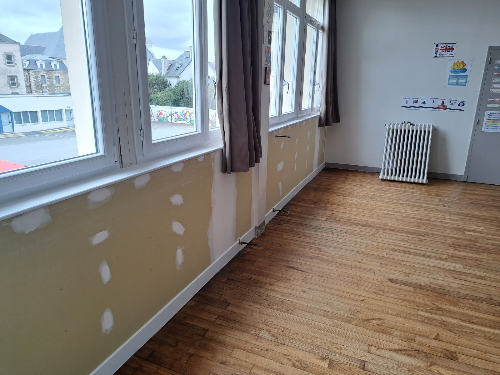 Intérieur d'une salle de classe vide ; sol en bois, fenêtres, murs beiges avec des zones rapiécées.