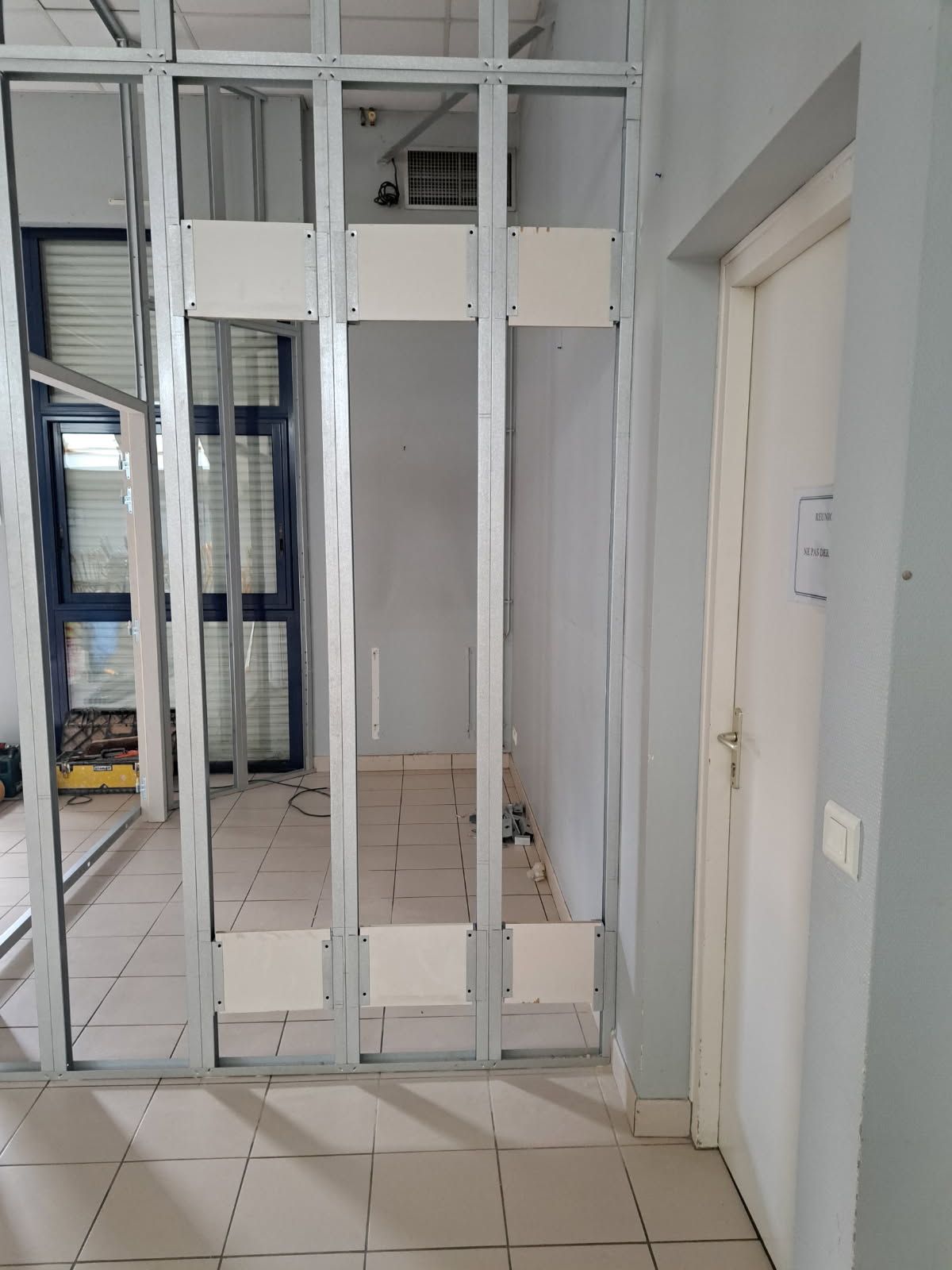 Pièce vide aux murs clairs, une porte, une fenêtre avec des stores et un petit escabeau.