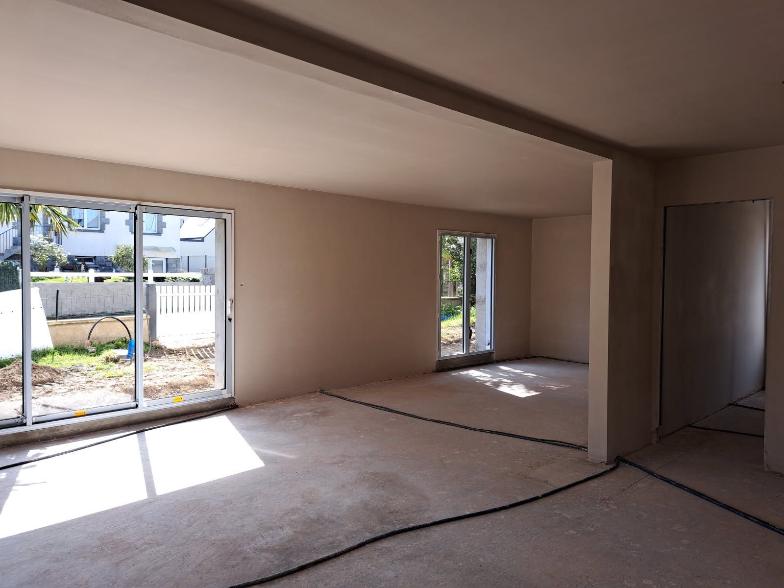 Pièce vide avec de grandes portes et fenêtres coulissantes en verre, des murs et un sol de couleur claire, et des câbles noirs courant au sol.