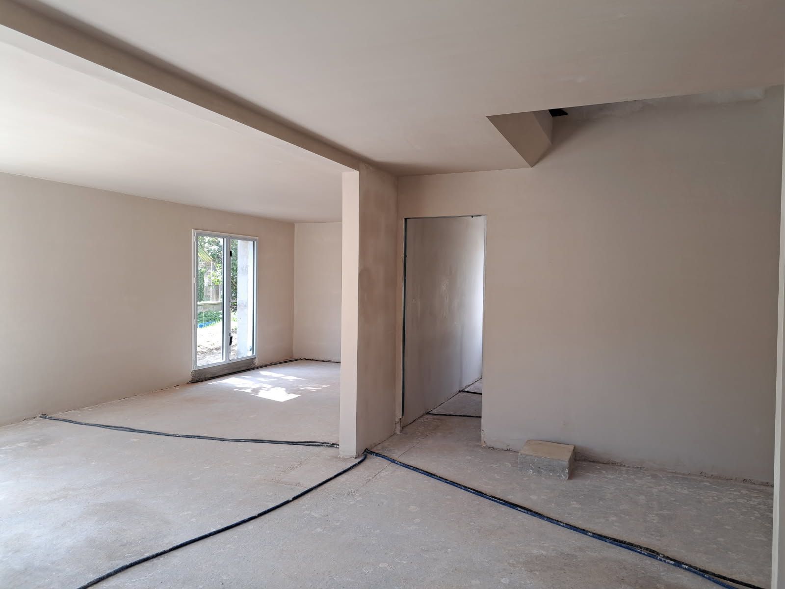 Intérieur d'une pièce en construction, avec des murs beiges, un sol non fini, des cadres de porte et des câbles apparents.